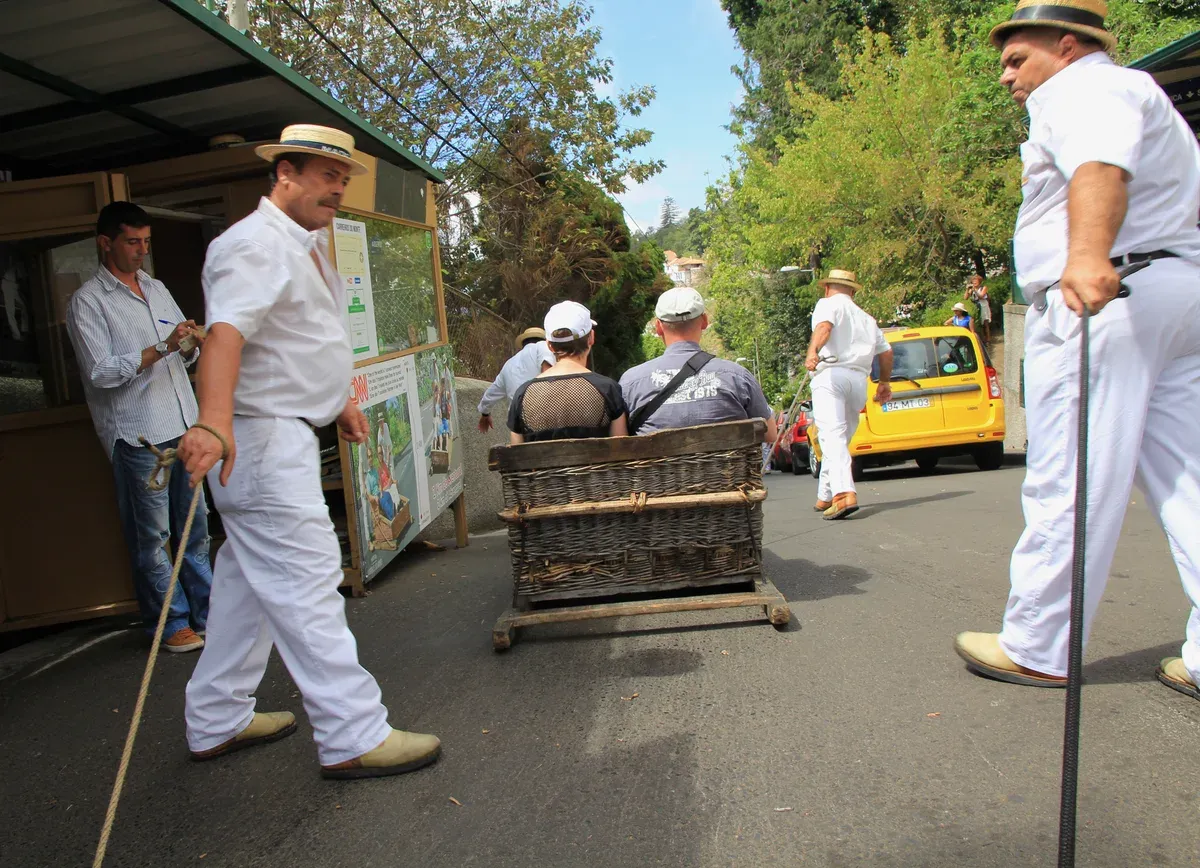 Two carreiros in white uniforms and straw hats steering a wicker basket toboggan down a paved street at the Monte departure point, Funchal, Madeira