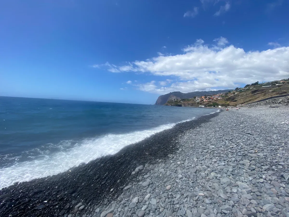 The long pebble and black sand shoreline of Praia Formosa beach in Funchal, Madeira, with the coastal hillside beyond