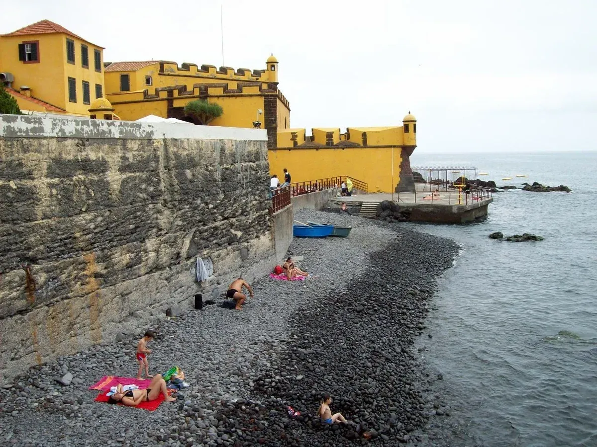 Sunbathers on the pebble Praia de São Tiago beach below the yellow fortress walls in Funchal, Madeira