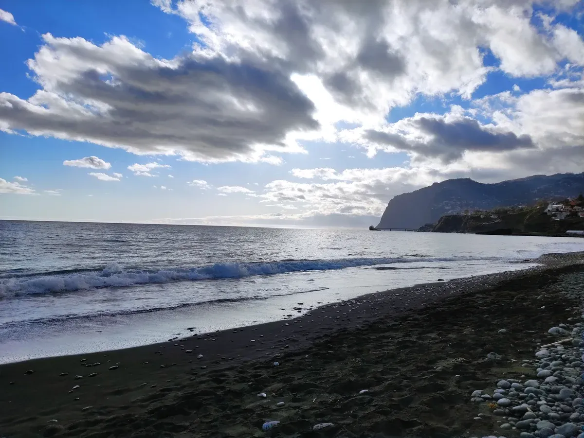 Waves rolling onto the black sand and pebble shore of Praia Formosa in Funchal, Portugal, with the coastline in the distance
